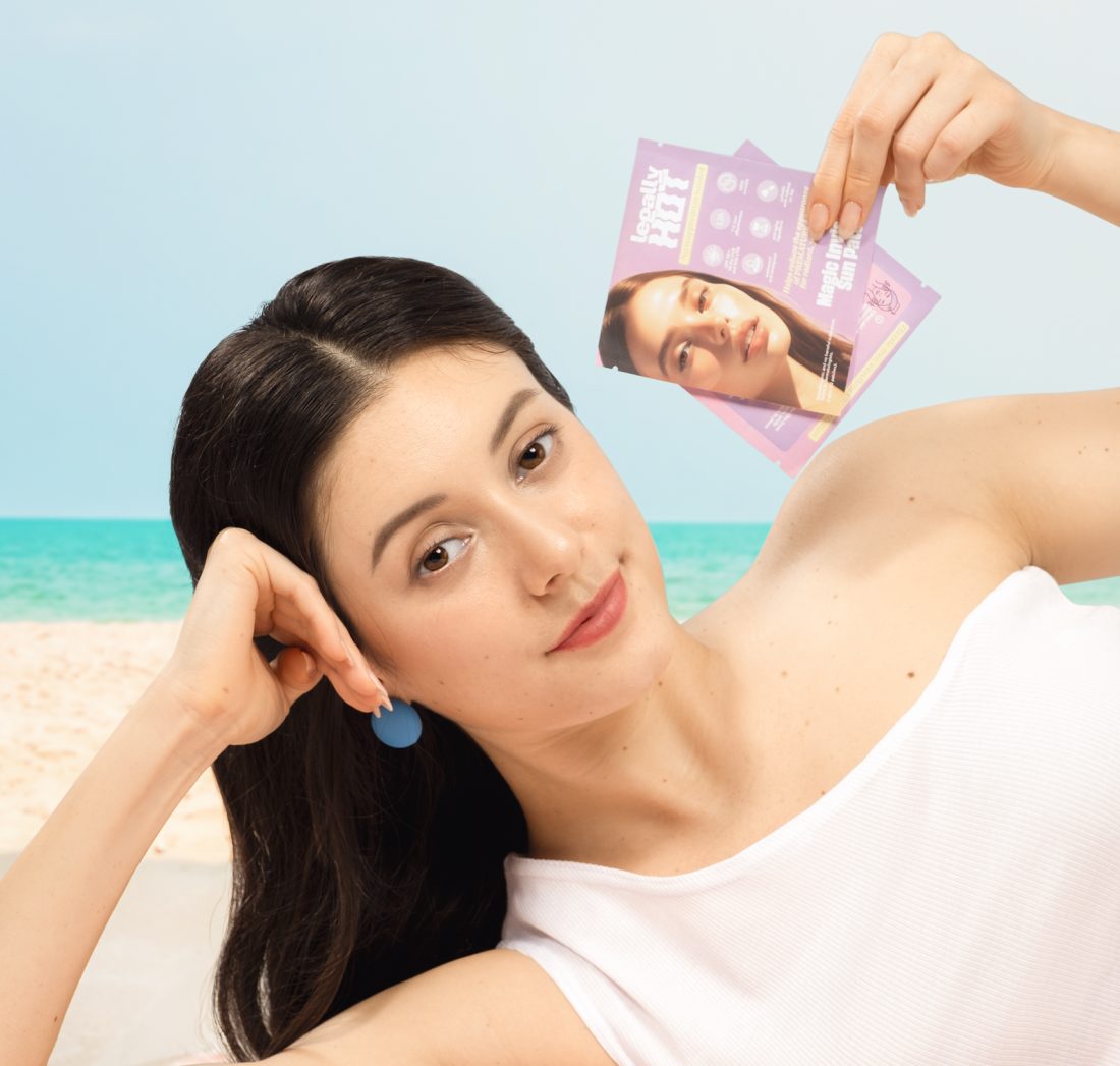 Woman on a beach towel holding Legally Hot Magic Invisible Sun Patch sachets